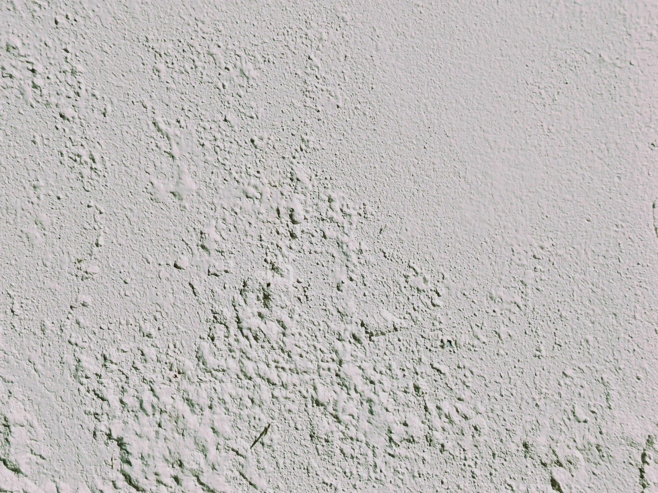 Close-up of a white textured wall with rough, uneven surfaces and small bumps, creating a tactile, stucco-like appearance. The lighting highlights the texture and shadows in the material.