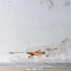 A close-up of a damaged wall with visible cracks and areas where plaster has chipped away, exposing underlying brickwork above a wooden baseboard.