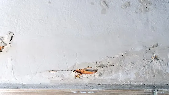 A close-up of a damaged wall with visible cracks and areas where plaster has chipped away, exposing underlying brickwork above a wooden baseboard.