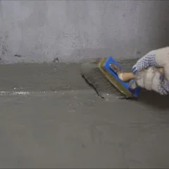 A person wearing a white work glove uses a brush to apply waterproofing sealant to a concrete floor and wall joint.