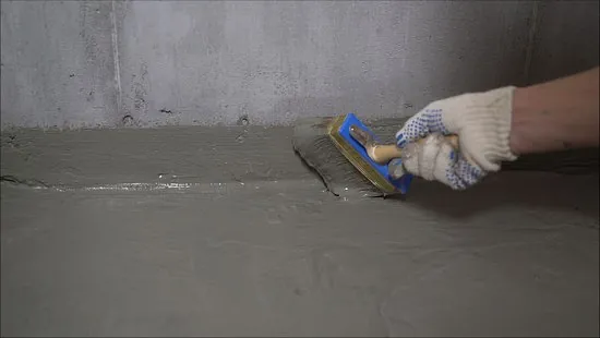A person wearing a white work glove uses a brush to apply waterproofing sealant to a concrete floor and wall joint.