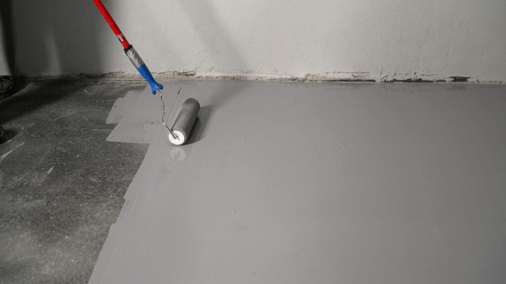 A paint roller with a red handle is being used to apply light gray paint to a concrete floor, partially covering the darker surface.