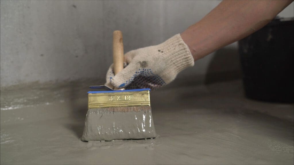 A gloved hand uses a large paintbrush to apply a thick, gray liquid coating onto a smooth concrete surface, likely as a waterproofing or sealing process.