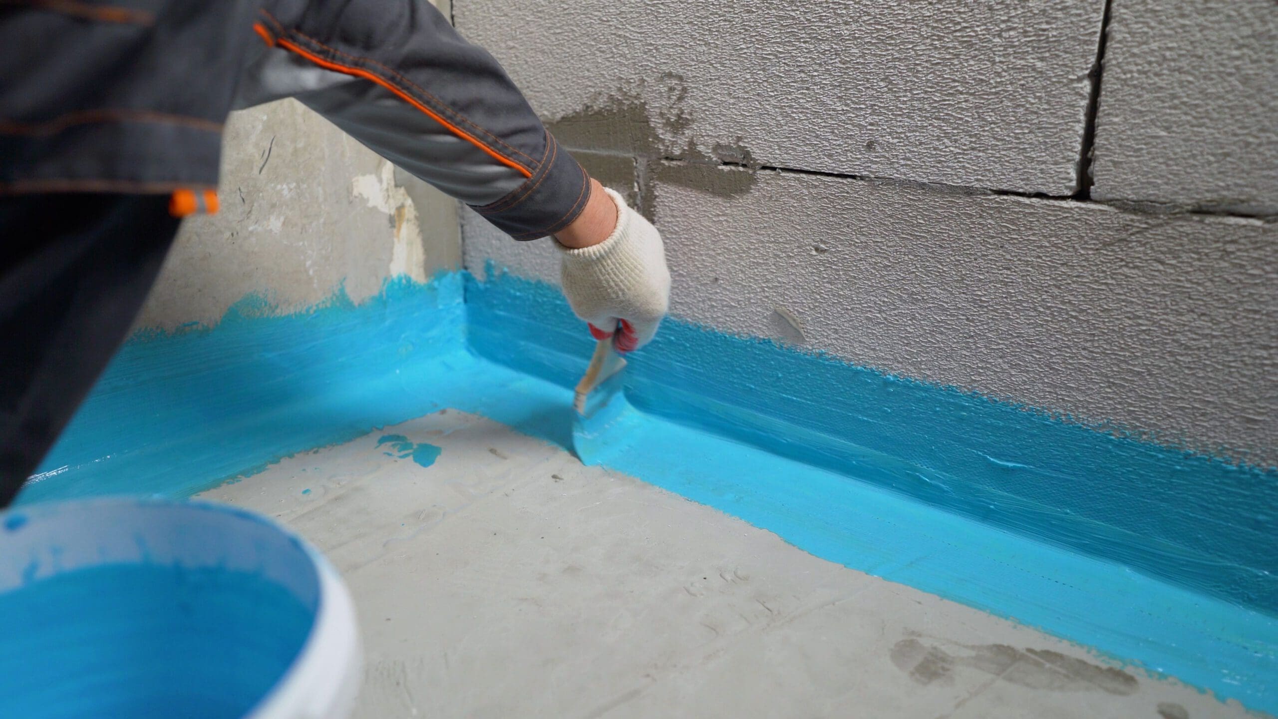 A person wearing a glove uses a brush to apply blue waterproofing paint along the corner seam between a concrete floor and wall. A bucket of blue paint sits nearby. The wall is unfinished with visible texture.