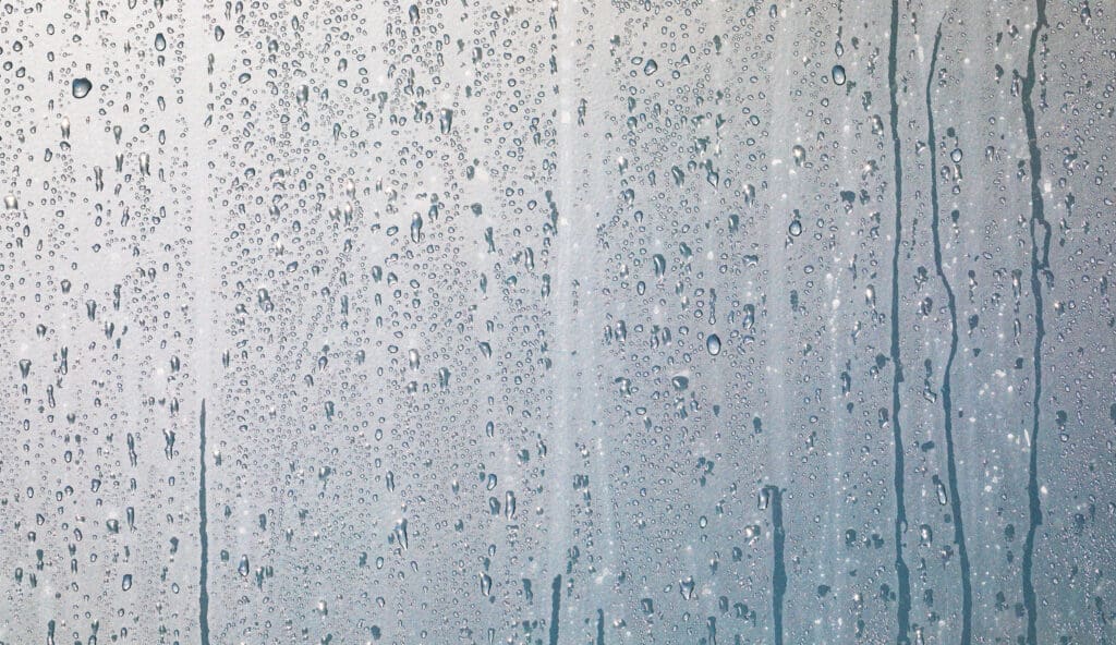 Water droplets and streaks on a foggy glass surface, suggesting rain or condensation, with a soft, diffused light in the background.