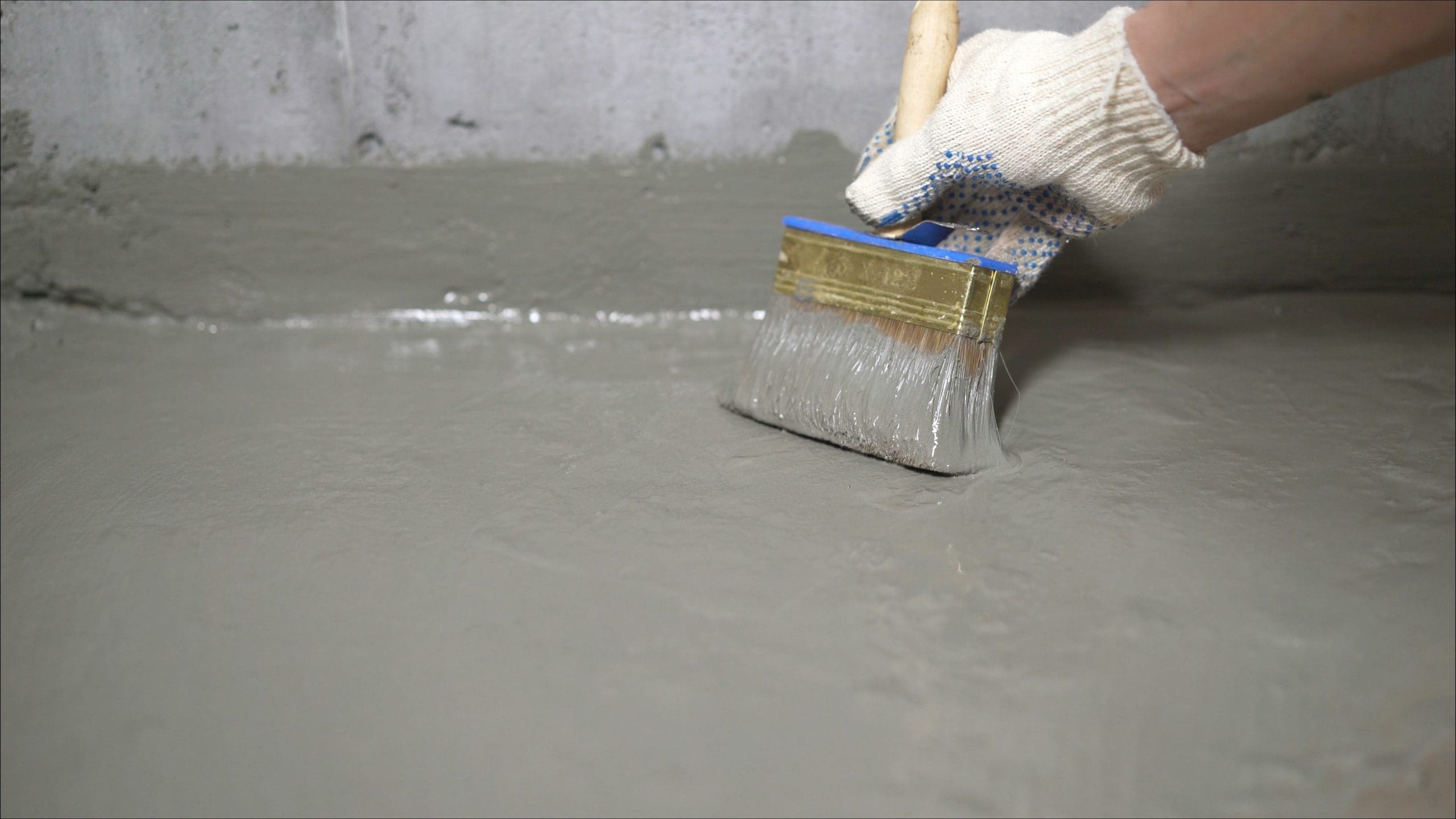 A gloved hand uses a wide brush to apply waterproofing sealant to a concrete surface, likely for construction or repair purposes.