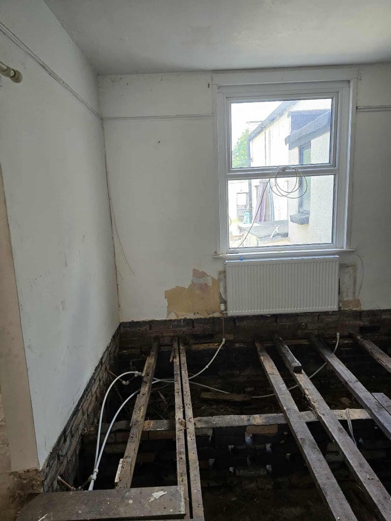 A room under renovation with floorboards removed, exposing wooden beams and wiring. The walls are partially damaged, and a window with a radiator underneath lets in natural light.