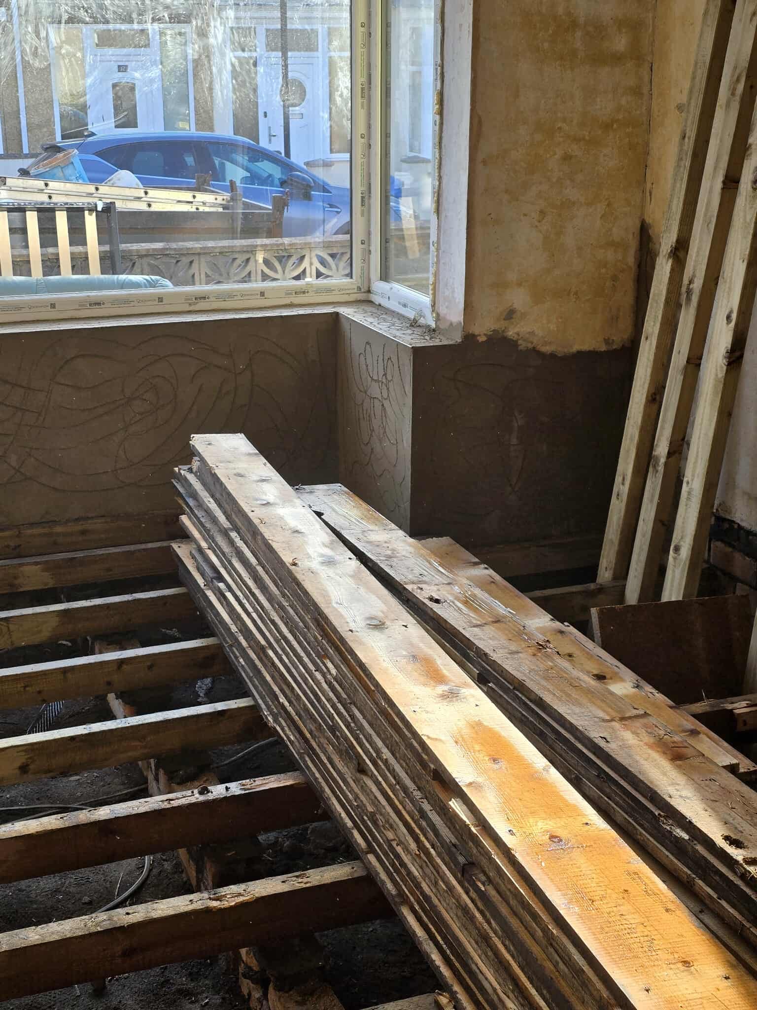 Stacks of wooden planks rest on exposed floor joists inside a room under renovation, with sunlight streaming through a large window and a car visible parked outside.