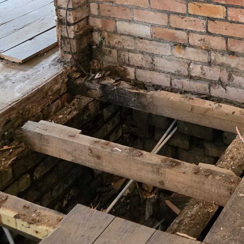 Exposed wooden beams and pipes under a damaged wooden floor next to a brick wall, showing part of a building’s subfloor structure under repair.